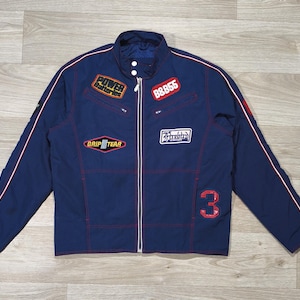 Peut inclure: Veste bleu marine avec des détails rouges et blancs. Elle comporte plusieurs écussons avec des textes et des chiffres, dont "POWER", "GRIP TEAR" et "88855". Le chiffre rouge "3" est en bas à droite. Fermeture éclair et col montant.