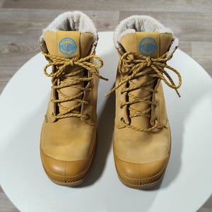 May include: Tan-coloured boots with light brown laces and a rubber toe cap. The boots feature a blue and white logo patch on the tongue and a soft, light-coloured lining. Designed for cold weather, these boots are suitable for various outdoor activities.