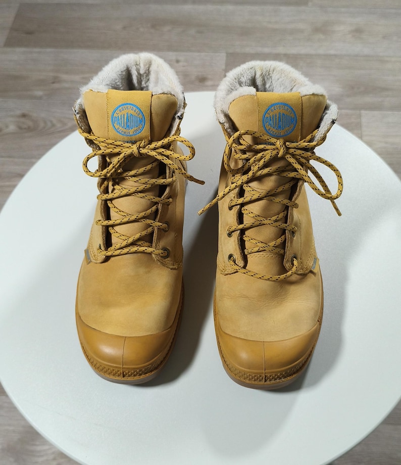 May include: Tan-coloured boots with light brown laces and a rubber toe cap. The boots feature a blue and white logo patch on the tongue and a soft, light-coloured lining. Designed for cold weather, these boots are suitable for various outdoor activities.