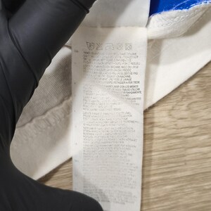 May include: Close-up of a white fabric tag with care instructions and text in multiple languages. The tag is attached to a white and blue garment. The text includes "PUUVILL" and "BUMBAC".