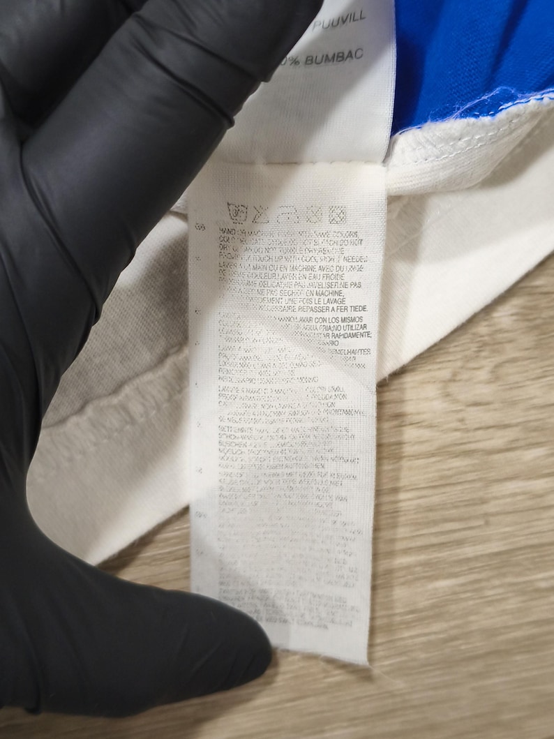 May include: Close-up of a white fabric tag with care instructions and text in multiple languages. The tag is attached to a white and blue garment. The text includes "PUUVILL" and "BUMBAC".