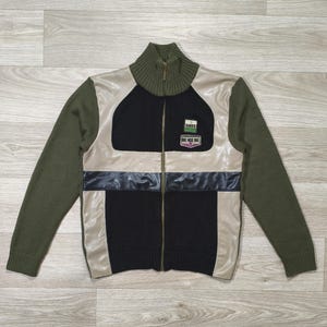 Peut inclure: Veste zippée avec des manches vert olive et un col montant. Le devant présente un motif à panneaux noirs et beiges avec une bande noire sur la poitrine. De petits écussons sont visibles sur le haut de la poitrine. La veste est faite de tricot et de similicuir.