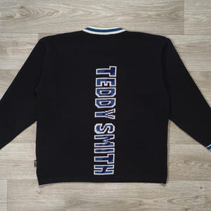 Peut inclure: Pull à manches longues noir avec le texte "TEDDY SMITH" verticalement dans le dos en bleu et blanc. Le pull a un col en V avec une bordure blanche et bleue, et une bordure bleue et blanche sur les poignets.