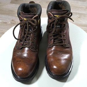 May include: Pair of brown leather work boots with laces. The boots have a padded collar and a black sole. The boots are designed for work and have a rugged appearance. The boots are shown on a white surface.