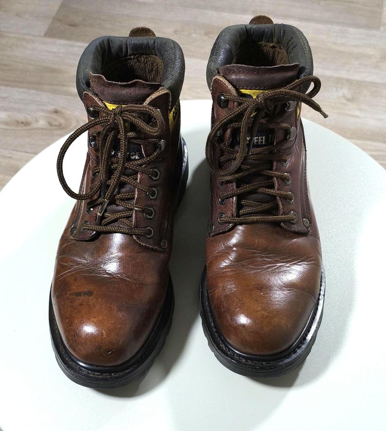 May include: Pair of brown leather work boots with laces. The boots have a padded collar and a black sole. The boots are designed for work and have a rugged appearance. The boots are shown on a white surface.