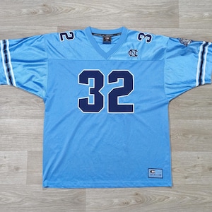 May include: Pale blue football jersey featuring the number 32 in navy blue on the front and shoulders. The jersey has a V-neck and white stripes on the sleeves. The University of North Carolina logo is visible on the chest.