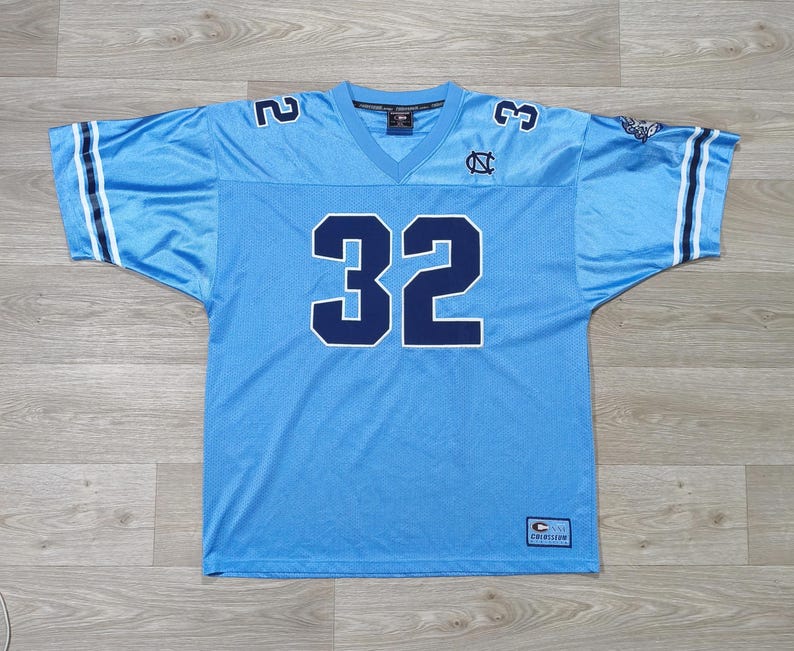 May include: Pale blue football jersey featuring the number 32 in navy blue on the front and shoulders. The jersey has a V-neck and white stripes on the sleeves. The University of North Carolina logo is visible on the chest.