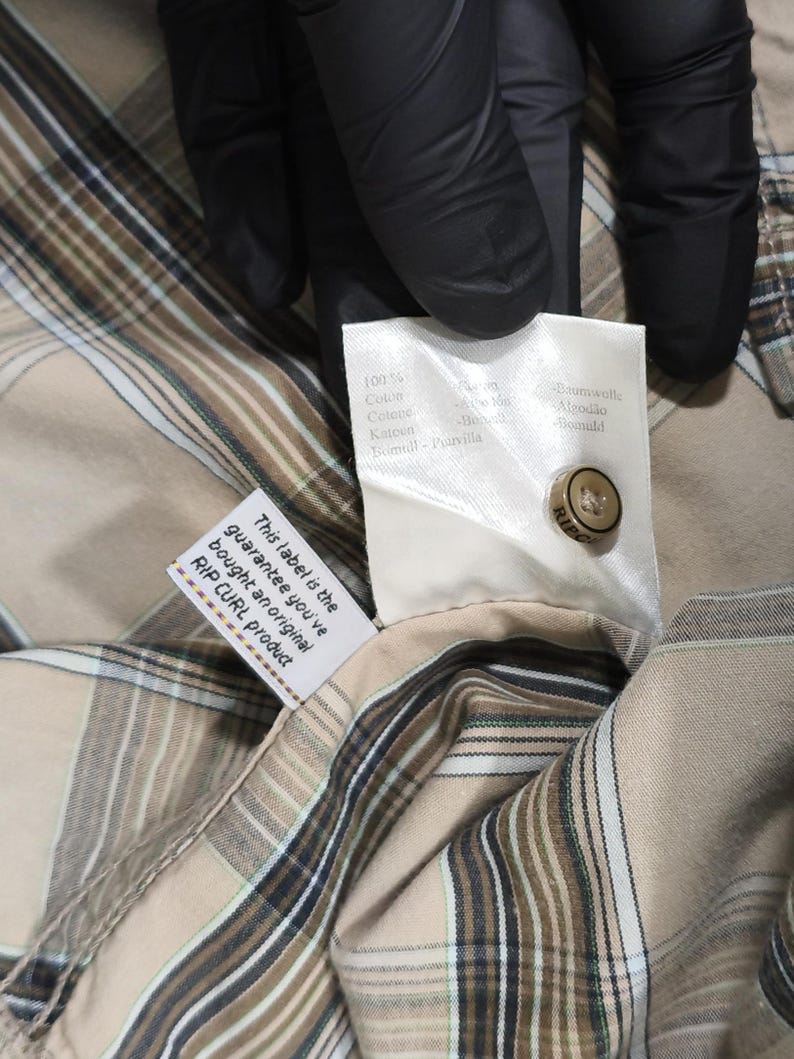 May include: A close-up shot of a tan and brown plaid shirt with a button and a tag. The tag reads "RIP CURL product" and "This label is the guarantee you've bought an original RIP CURL product." The shirt has a classic plaid pattern and is made of a soft fabric.