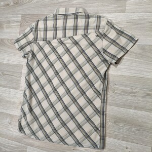 May include: Short-sleeved shirt with a plaid pattern in beige, grey, and blue. The shirt has a collar and a small logo on the back. The plaid design is diagonal across the front.