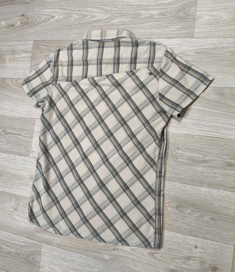 May include: Short-sleeved shirt with a plaid pattern in beige, grey, and blue. The shirt has a collar and a small logo on the back. The plaid design is diagonal across the front.
