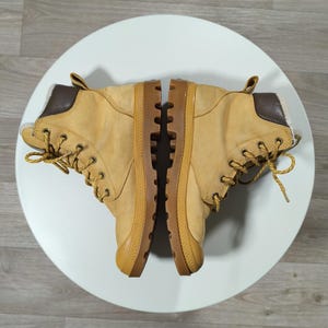 May include: A pair of tan boots with brown details and laces. The boots have a robust design with a thick sole and are placed on a white surface. The boots appear to be made of leather or a similar material.