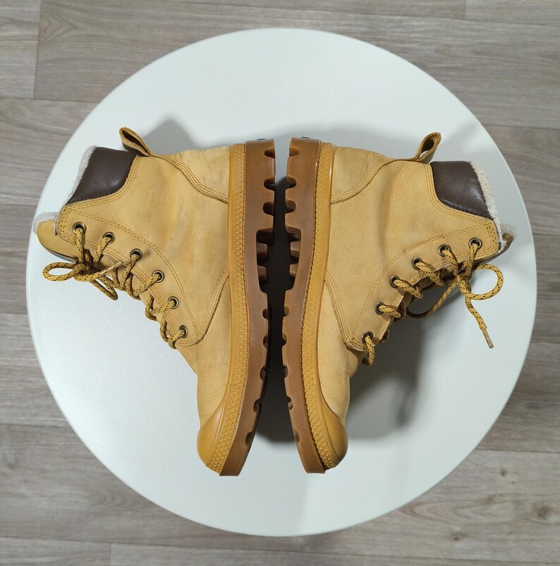 May include: A pair of tan boots with brown details and laces. The boots have a robust design with a thick sole and are placed on a white surface. The boots appear to be made of leather or a similar material.