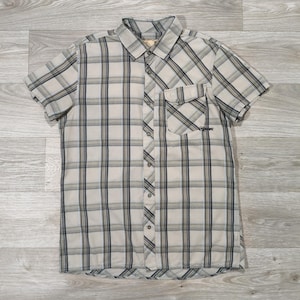May include: A short-sleeved shirt with a plaid design in beige, navy, and light blue. The shirt features a button-down front, a collar, and a chest pocket. The garment is displayed flat on a wooden surface.