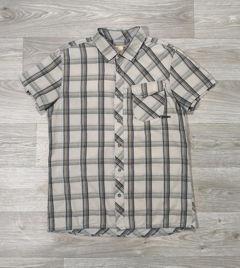 May include: A short-sleeved shirt with a plaid design in beige, navy, and light blue. The shirt features a button-down front, a collar, and a chest pocket. The garment is displayed flat on a wooden surface.