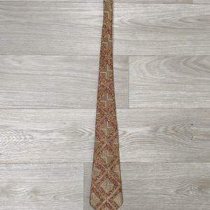 Könnte beinhalten: Eine braune Krawatte mit einem sich wiederholenden geometrischen Muster aus roten und goldenen Rauten. Die Krawatte besteht aus glänzendem Material und wird auf einer hellen Holzoberfläche präsentiert. Ein klassisches Accessoire.