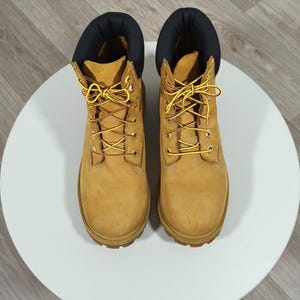 Puede incluir: Un par de botas de cuero color canela con cuello acolchado negro, ojales de metal dorado y cordones estampados en amarillo y marrón. Las botas tienen una puntera redondeada y una suela gruesa y duradera para un calzado de exterior.