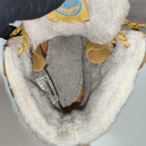 May include: Close-up of the interior of a tan and cream-coloured boot. The boot has a soft, fuzzy lining and a yellow tag with blue text. The laces are yellow with black dots. The boot is open, revealing the inside.
