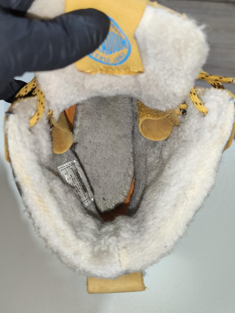 May include: Close-up of the interior of a tan and cream-coloured boot. The boot has a soft, fuzzy lining and a yellow tag with blue text. The laces are yellow with black dots. The boot is open, revealing the inside.