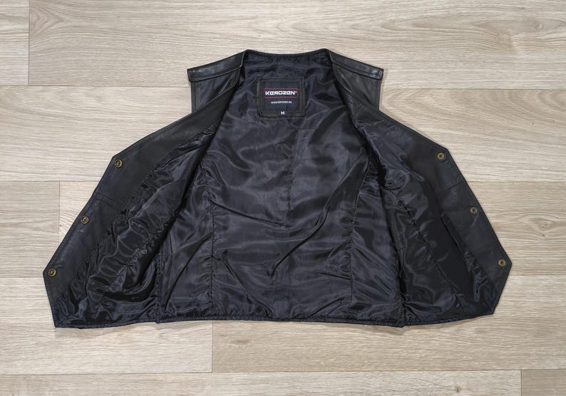 May include: Black leather vest with a satin lining, open to display the interior. The vest has a V-neck, button closures, and a label that reads "KONDORON". The vest is laid flat on a light wood grain surface.