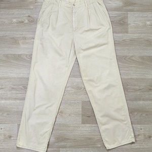 May include: A pair of cream-colored trousers featuring a high-waisted design with double front pleats, a button and zipper fly closure, side pockets, and a straight-leg silhouette. The pants are constructed from a solid, light-toned fabric.