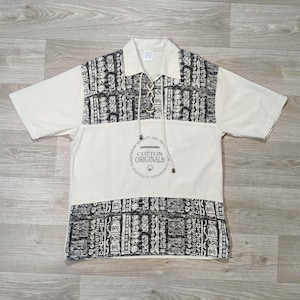 Peut inclure: Chemise crème à manches courtes avec un col et un motif noir et blanc. La chemise présente une fermeture à cordon et le texte "COTTON ORIGINALS" dans un emblème circulaire.
