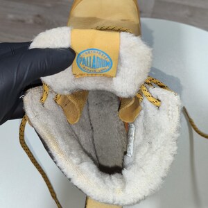 May include: Tan winter boot with a cream-coloured, fuzzy interior. The boot features a tan leather exterior, yellow laces, and a blue and yellow label with the text "PALLADIUM".