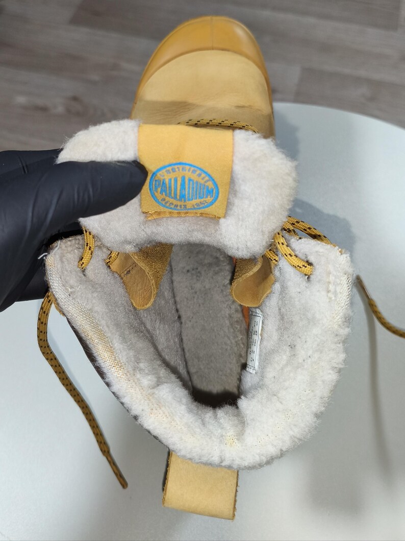 May include: Tan winter boot with a cream-coloured, fuzzy interior. The boot features a tan leather exterior, yellow laces, and a blue and yellow label with the text "PALLADIUM".