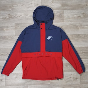 Peut inclure: Veste anorak Nike Air bleu marine et rouge avec capuche. Le logo Nike Air blanc est sur la poitrine et l'inscription "NIKE AIR" est sur le devant. La veste a un cordon de serrage en bas.
