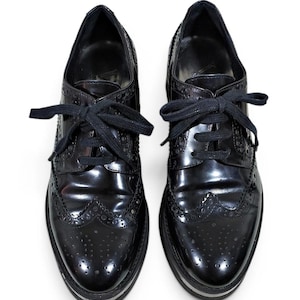 May include: A pair of black patent leather oxford shoes featuring classic brogue detailing, wingtip toes, and black laces. The shoes have a thick, layered sole with a visible silver-toned stripe running along the side of the platform base.