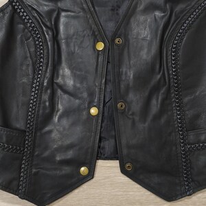 May include: Black leather vest with a V-neck and gold-colored buttons. The vest features decorative braided trim along the edges and pockets. The vest is a classic style.