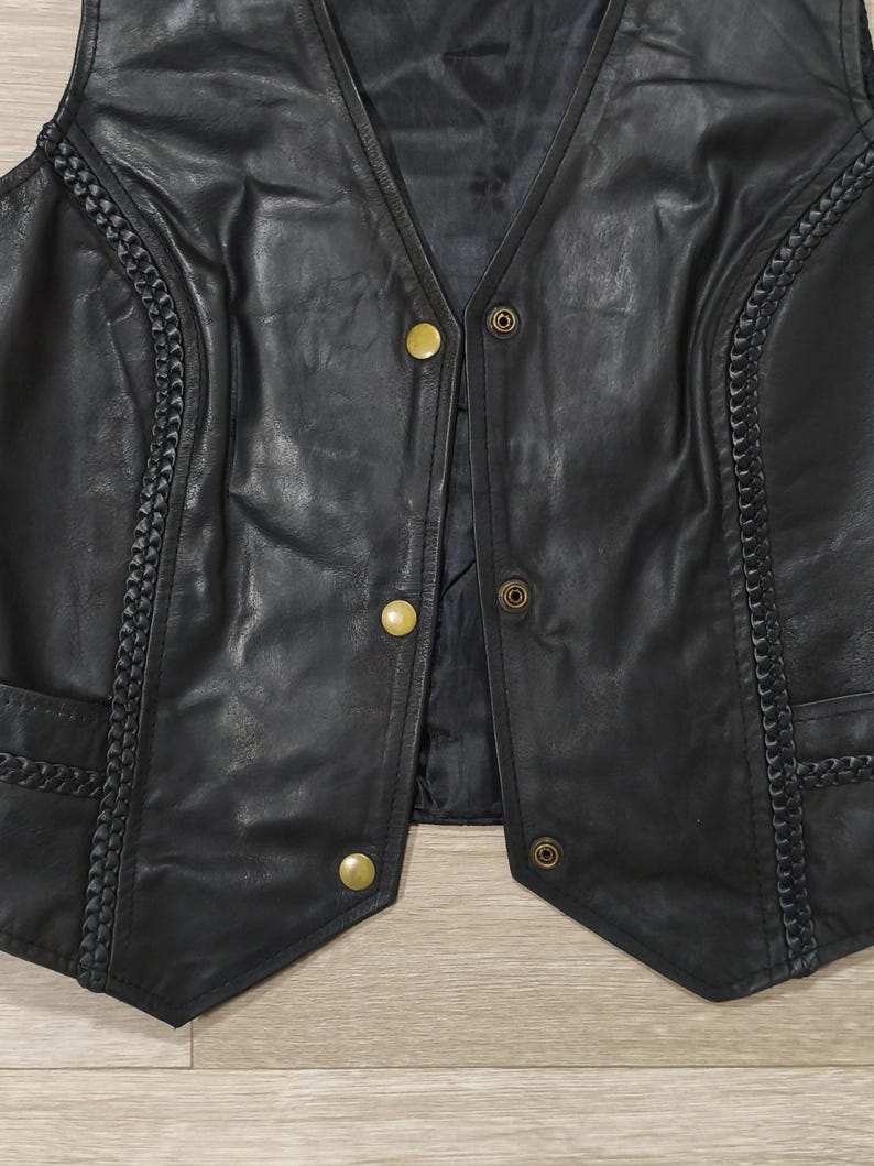 May include: Black leather vest with a V-neck and gold-colored buttons. The vest features decorative braided trim along the edges and pockets. The vest is a classic style.