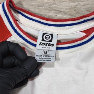 May include: White and red athletic shirt with blue and red trim around the collar. The shirt has a tag that reads "lotto" and "Italian Sport Design". The shirt is a size medium.