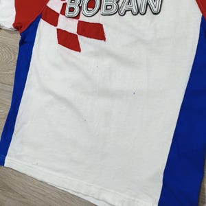 May include: White t-shirt with red and blue accents. The shirt features the word "BOBAN" in black outlined letters, and a red and white checkered design. The sleeves are red and the sides have blue panels.