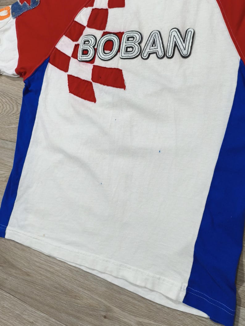May include: White t-shirt with red and blue accents. The shirt features the word "BOBAN" in black outlined letters, and a red and white checkered design. The sleeves are red and the sides have blue panels.