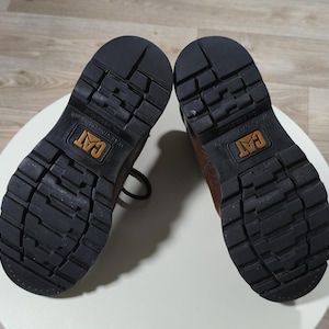 May include: Pair of brown leather work boots with black rubber soles. The soles have a deep tread pattern for traction and the CAT logo is visible on the sole. The boots are shown from the bottom.