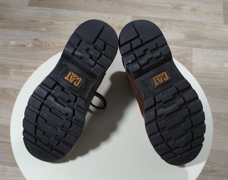 May include: Pair of brown leather work boots with black rubber soles. The soles have a deep tread pattern for traction and the CAT logo is visible on the sole. The boots are shown from the bottom.