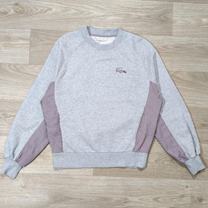 Könnte beinhalten: Ein hellgraues Sweatshirt mit Rundhalsausschnitt, langen Raglanärmeln und einem kleinen gestickten Krokodil-Logo auf der linken Brust. Das Kleidungsstück hat kontrastierende Seiteneinsätze mit einem dunkelroten und weißen Karomuster sowie gerippte Bündchen.