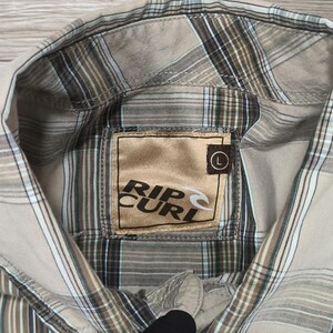 May include: A beige and brown checked shirt with the "Rip Curl" logo on the inside collar. The shirt features a classic collar and button closure. The shirt is a size large.