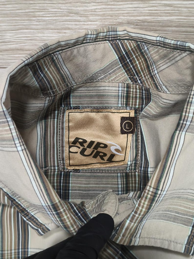 May include: A beige and brown checked shirt with the "Rip Curl" logo on the inside collar. The shirt features a classic collar and button closure. The shirt is a size large.