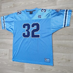 May include: Light blue football jersey with the number 32 in navy blue. The jersey has a V-neck and the number 2 and 3 on the shoulders. The sleeves have white and navy blue stripes. The jersey has the logo of the University of North Carolina.