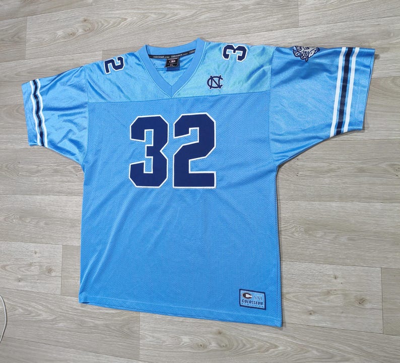 May include: Light blue football jersey with the number 32 in navy blue. The jersey has a V-neck and the number 2 and 3 on the shoulders. The sleeves have white and navy blue stripes. The jersey has the logo of the University of North Carolina.