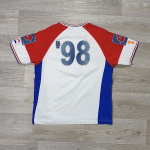 May include: White, red, and blue short-sleeved t-shirt with the number '98' in denim-style fabric on the back. The shirt has a color block design with red sleeves and blue side panels. The collar and sleeve cuffs have red and blue stripes.