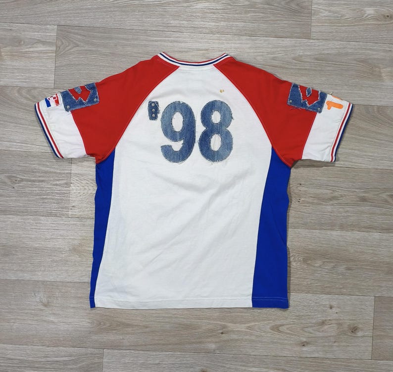 May include: White, red, and blue short-sleeved t-shirt with the number '98' in denim-style fabric on the back. The shirt has a color block design with red sleeves and blue side panels. The collar and sleeve cuffs have red and blue stripes.
