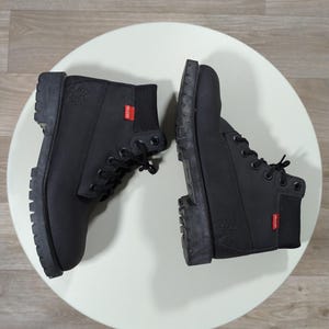 May include: A pair of black work boots with chunky soles and black laces, presented on a white surface. Each boot features a red tag with white text. The boots are constructed from a robust, woven fabric.