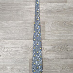 Peut inclure: Une cravate grise avec un motif répété de petites figures tenant des panneaux jaunes et blancs. Les figures ont des accents bleus et blancs. La cravate est en tissu lisse, présentée sur une surface en bois clair.