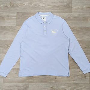 May include: A light blue long-sleeve polo shirt. The shirt features a collared neckline with a three-button placket and a white embroidered logo on the chest. The shirt has the text "LACOSTE 1927" embroidered on the chest.