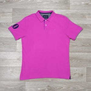 Peut inclure: Un polo magenta vif à manches courtes. Le polo a un col classique, une patte de boutonnage à trois boutons et un petit logo près de la poitrine. La manche gauche porte le chiffre "0" bleu marine. Le tissu est doux et respirant.
