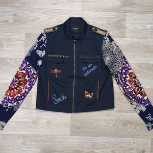 Peut inclure: Une veste zippée bleu marine avec des manches longues ornées de motifs floraux et mandalas complexes en rouge, violet et blanc. Le devant présente deux poches poitrine, des épaulettes dorées et des broderies indiquant Smile et We are animals.