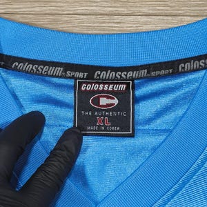 May include: A close-up of a light blue Colosseum Sport XL jersey. The jersey has a V-neck and a tag that reads "THE AUTHENTIC" and "MADE IN KOREA". The Colosseum logo is visible on the tag and the collar.