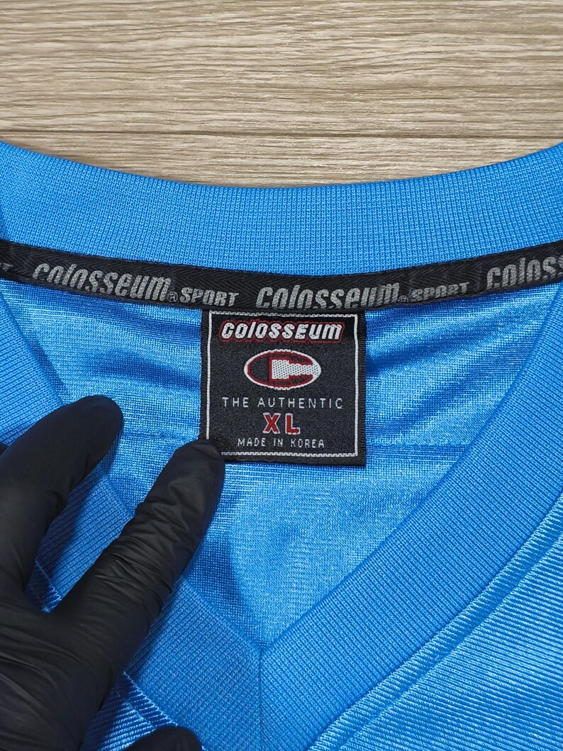 May include: A close-up of a light blue Colosseum Sport XL jersey. The jersey has a V-neck and a tag that reads "THE AUTHENTIC" and "MADE IN KOREA". The Colosseum logo is visible on the tag and the collar.